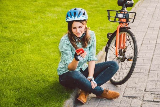 A young cyclist sitting on the ground next to her orange bike, checking her phone after making cycling mistakes.