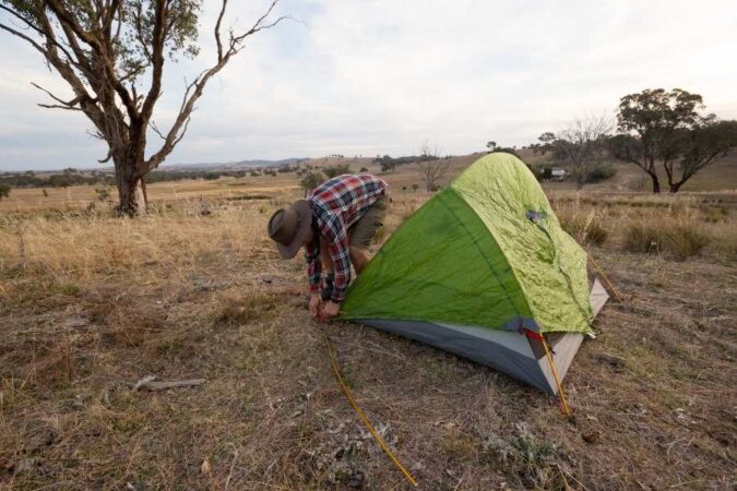 A camper sets up a green waterproof camping tent in a dry open field during the late afternoon.